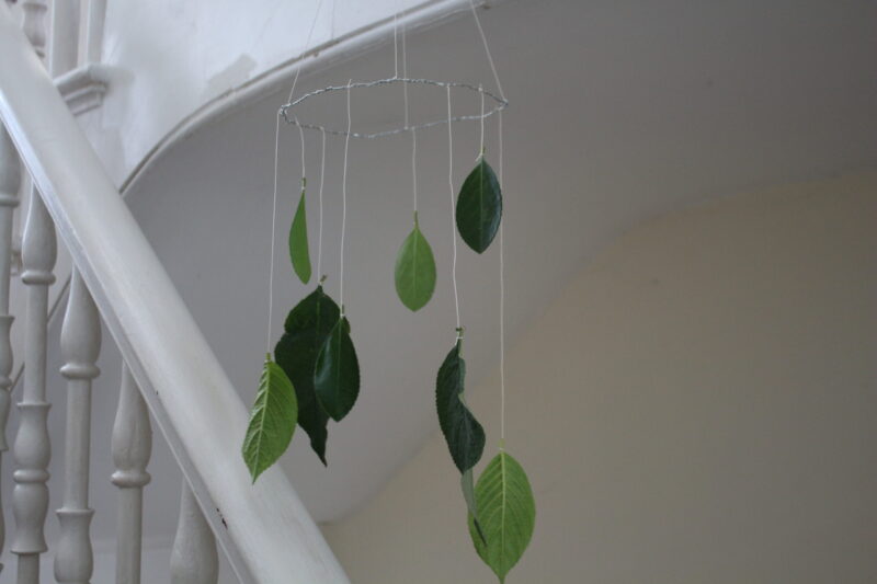A photograph of a mobile made from silver metal wire and green oval-shaped leaves, hanging in the middle of a white staircase.
