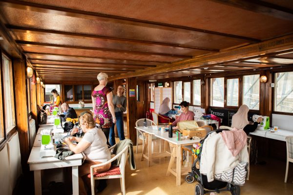 A photograph of the Dresscue space on ONCA Barge, people are sat at tables working on sewing machines whilst others stand trying on their creations or chatting together.