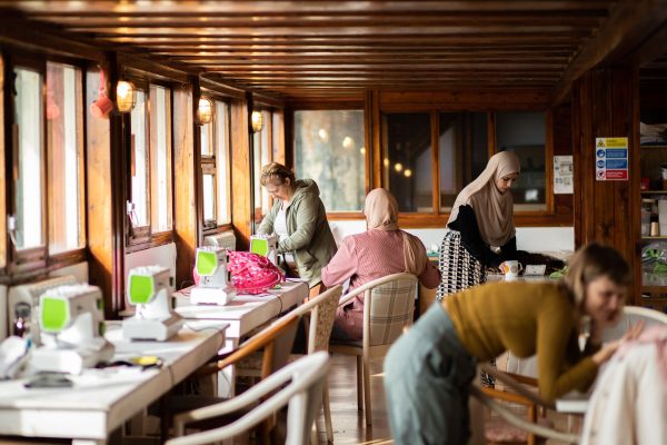 A photograph of people in the Dresscue sewing space on ONCA Barge. To the left are table lined up with sewing machines and people working on creative projects.